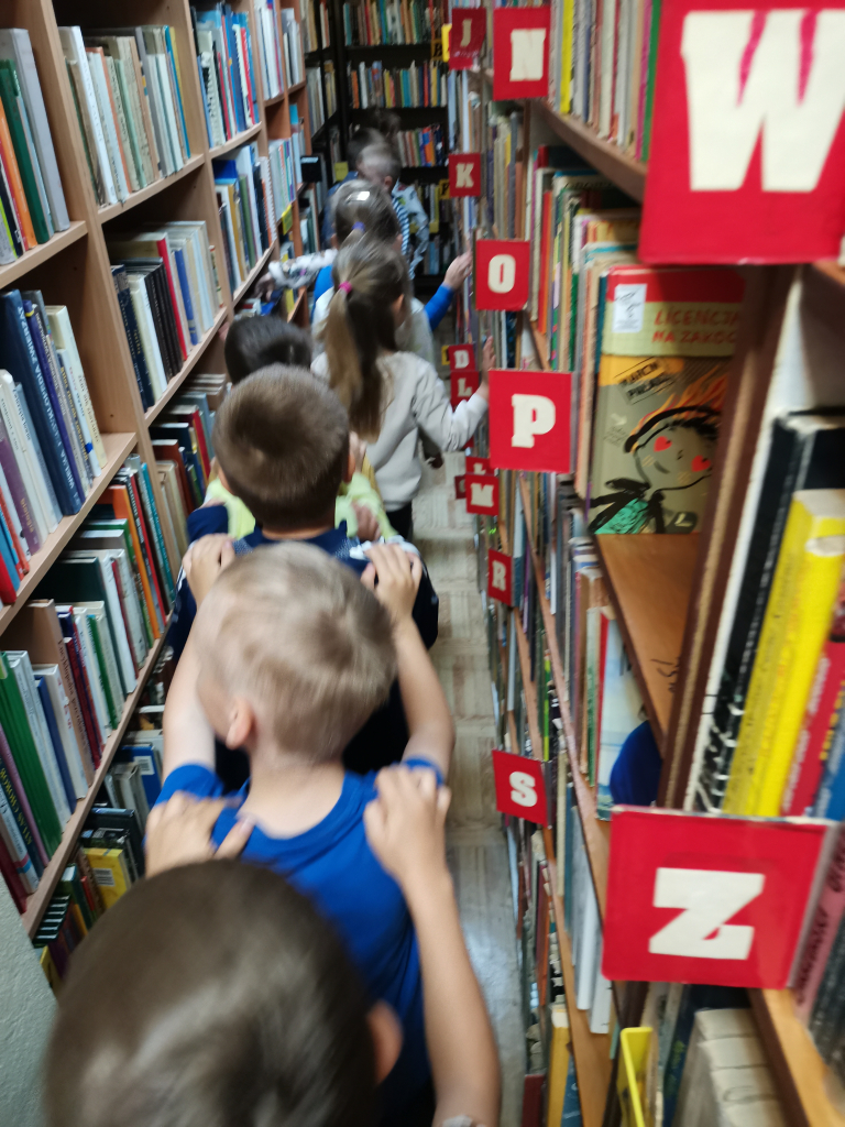 Zdjęcie przedstawia grupę pięcio i sześciolatków zwiedzających bibliotekę bawiąc się w zabawę Wesoły pociąg