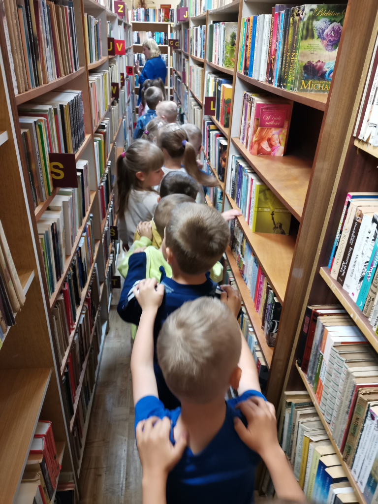 Zdjęcie przedstawia grupę przedszkolaków zwiedzających bibliotekę bawiąc się w zabawę Wesoły pociąg