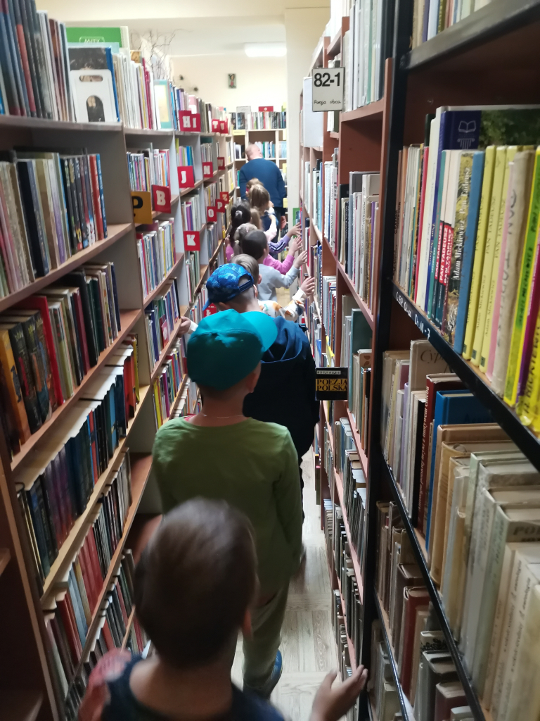 Zdjęcie przedstawia grupę przedszkolaków zwiedzających bibliotekę bawiąc się w zabawę Wesoły pociąg