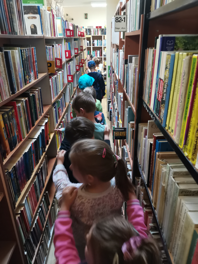 Zdjęcie przedstawia grupę sześciolatków zwiedzających bibliotekę bawiąc się w zabawę Wesoły pociąg