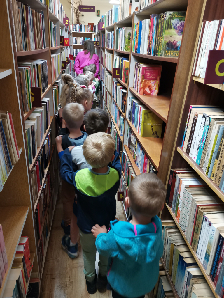 Zdjęcie przedstawia grupę pięcio i sześciolatków zwiedzających bibliotekę bawiąc się w zabawę Wesoły pociąg